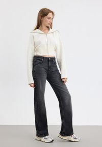 Gekürzte weiße Zip-Hoodie mit weiten Ärmel, kombiniert mit dunkelgrauen High-Waist-Jeans und mehrfarbigen klobigen Sneakers. Flacher Hintergrund.