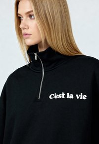 Sweat-shirt noir à fermeture éclair avec col haut, arborant l'inscription blanche « C'est la vie » sur le devant, fabriqué en tissu doux à la texture lisse.