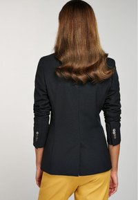 Blazer noir ajusté avec des poignets à boutons et un design ajusté, doté d'un tissu texturé et d'une fente à l'arrière.