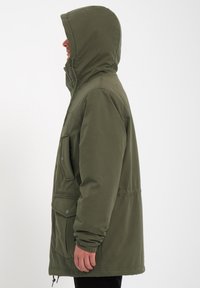Parka de color verde oliva con capucha, que cuenta con bolsillos delanteros con botones a presión, cierre con cremallera y un corte holgado. Fabricada en tejido texturizado.
