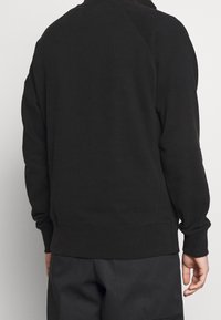 Persoon in een eenvoudige zwarte hoodie en donkere broek, van achteren gezien tegen een effen witte achtergrond.