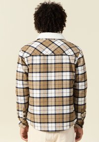 Camicia in flanella a quadri marrone con accenti bianchi e blu navy, dotata di un colletto foderato in sherpa e polsini con bottoni, mostrata dal retro.