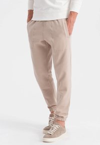 Beige Jogginghosen aus weichem Stoff, mit Seitentaschen, elastischen Knöchelbündchen und dekorativen Nähdetails, kombiniert mit grauen Sneakers.