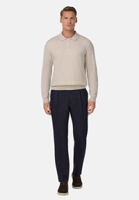 Gai beigā adīta pulovere ar polo apkakli, savienots ar tumši ziliem krokotiem biksēm un brūniem apaviem. Gluda tekstūra un brīva piegriezuma.