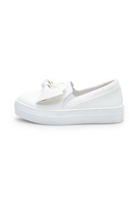 Fehér slip-on sportcipők, texturált gumitalppal, sima szintetikus felső résszel és egy nagy masni díszítéssel elöl.