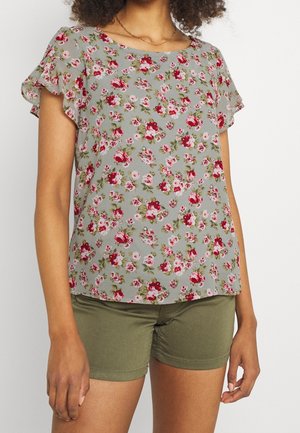 Kobieta w krótkim rękawie, szarej bluzce w kwiaty z czerwonymi i różowymi kwiatami, w połączeniu z oliwkowozielonymi szortami.
