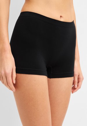 Femme portant un short noir uni et ajusté, debout, les mains détendues le long du corps sur un fond clair et uni.