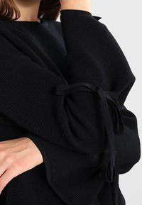 Svart överdimensionerad topp med ribbad textur, med vida ärmar som är bundna med tygremsor vid ärmsluten. Materialet verkar mjukt och lätt.