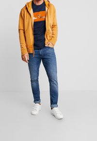 Sudadera amarilla con cremallera y bolsillos frontales, camiseta azul marino con texto naranja, jeans azules y zapatillas blancas. Conjunto casual sobre un fondo neutro.