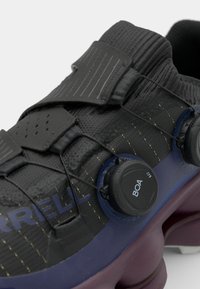 Chaussure de sport noire avec un empeigne texturée, équipée d'un système de laçage BOA, une semelle intermédiaire violette et des détails de couture jaunes.