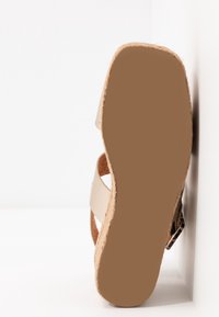 Sandale beige avec deux brides en cuir croisées et une semelle en liège, inclinée pour montrer la base courbée et la texture des matériaux.