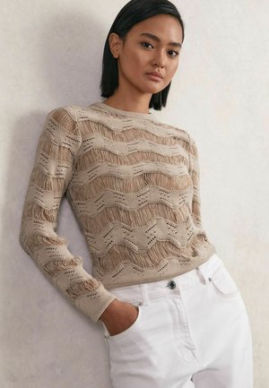 Suéter de punto beige con un patrón de olas y textura de flecos, combinado con pantalones de mezclilla blancos. La modelo posa con las manos en los bolsillos.