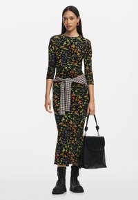 Fekete virágmintás ruha narancs, lila és zöld nyomattal. Hosszú ujjú, testhezálló sziluett, a derekán megkötött kockás inggel. Fekete kézitáska és csizma.