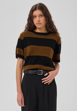 Femme aux longs cheveux bouclés porte un pull rayé marron et noir à manches courtes et un pantalon noir avec une ceinture noire, debout devant un fond uni.
