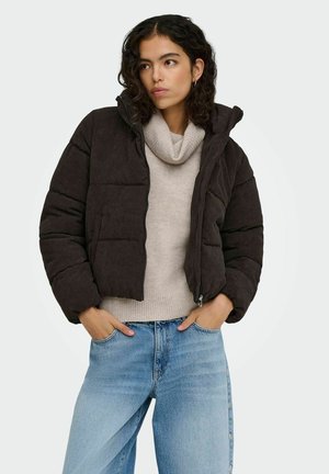 Femme portant une doudoune marron foncé par-dessus un pull beige à col bénitier et un jean bleu clair, debout les mains dans les poches.