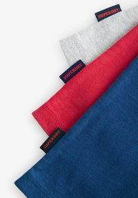 Tres camisetas de algodón apiladas: gris, roja y azul. Cada una presenta una etiqueta de logo visible con "SUPERDRY" en color naranja. Diseño simple y sencillo.