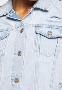 Světle modrá džínová bunda s klasickým designem, kovovými knoflíky a opotřebenými detaily na ramenou a kapsách.