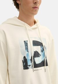 Sudadera crema con un gráfico de montañas y nubes, con un diseño de letras grandes en negro, cordones y una textura suave.