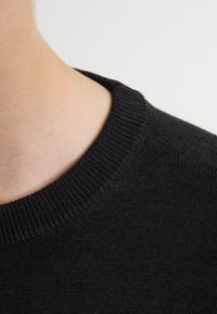 Maglione nero a coste con scollo rotondo, che mette in evidenza un tessuto testurizzato. Primo piano della zona del collo e della spalla.