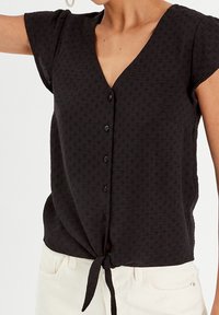 Haut noir à manches courtes avec encolure en V, boutons à l'avant et ceinture nouée à la taille. Tissu texturé avec des détails de motifs subtils, assorti à un pantalon clair.