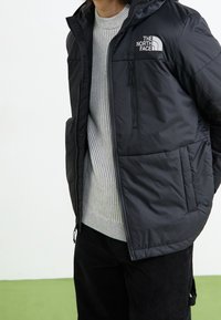 Černá zateplená bunda s kapucí, která má zipovou kapsu, logo The North Face na hrudi a hladký, lesklý materiál.