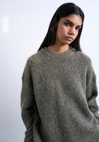 Jeune femme aux longs cheveux bruns portant un pull en maille marron-gris duveteux, debout devant un fond uni gris clair.