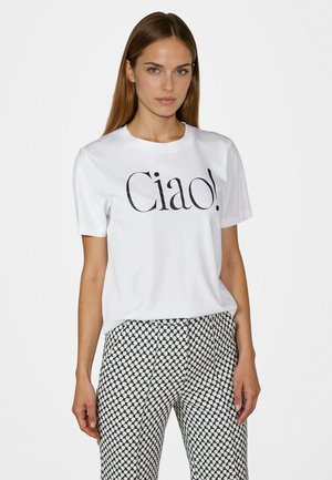Frau mit langen hellbraunen Haaren, die ein weißes T-Shirt mit dem Text "Ciao!" und schwarz-weiß gemusterte Hosen trägt, steht vor einem einfarbigen Hintergrund.