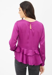 Blusa de satén fucsia con mangas largas, dobladillo estilo peplum, detalle de ojal en la espalda y textura suave; combinada con jeans oscuros.