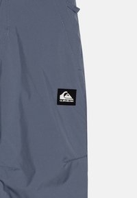 Grå vattenavvisande jacka med synliga sömmar och en Quiksilver-logotyp. Har en dragkedjeficka och justerbara manschetter. Slät textur.