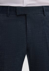 Marineblauwe broek van een gestructureerde stof, met een platte voorkant en lusjes voor een riem. Het ontwerp benadrukt een getailleerde pasvorm en een subtiel patroon.