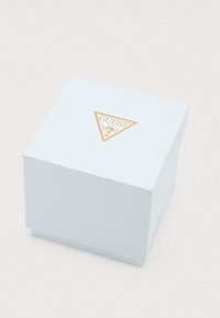 Λευκό τετράγωνο κουτί με υφή, με χρυσό τριγωνικό λογότυπο Guess και κείμενο "Guess U.S.A.? Washed Jeans" στο καπάκι, πάνω σε απλό φόντο.