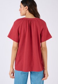 Top rosso strutturato con maniche corte e dettaglio arricciato sul retro, abbinato a pantaloni azzurri chiari. Sfondo bianco semplice.
