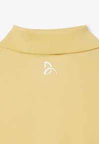 Prenda amarilla con un cuello alto acanalado y un pequeño logo minimalista blanco centrado debajo del escote.