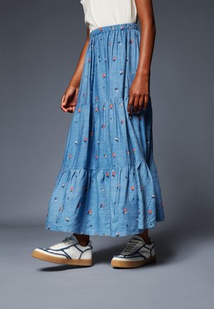 Persona que lleva una falda de volantes azul con estampado floral, camiseta blanca y zapatillas blancas con detalles azules, de pie frente a un fondo gris liso.