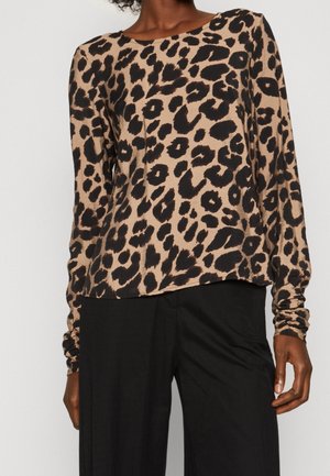 Personne portant un haut à manches longues beige avec un imprimé léopard noir et un pantalon large noir, sur un fond clair uni.