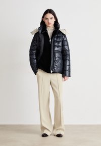 Schwarze Puffjacke mit einer beigen Kapuze, Reißverschluss vorne und zwei Seitentaschen. Kombiniert mit einem schwarzen Pullover und cremefarbigen weit geschnittenen Hosen.