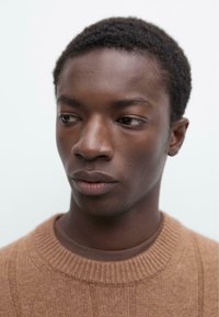 Brauner gerippter Pullover an einer Person mit kurzen, lockigen Haaren und glatter Haut. Der Hintergrund ist in einem hellen, neutralen Farbton gehalten.