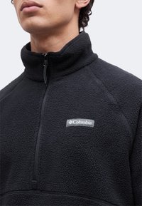 Άνδρας που φοράει μαύρο υφασμάτινο μπουφάν fleece με φερμουάρ μέχρι τη μέση και εμφανές λογότυπο Columbia στο στήθος.