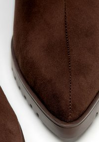 Botas de ante marrón hasta el tobillo con punta afilada, que presentan un detalle de costura única y una suela de goma texturizada. Acabado suave con un diseño minimalista.