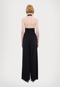 Svart halter jumpsuit med et tettsittende liv, åpen rygg og vide ben. Glatt stoff med glidelås bak.