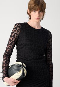 Schwarzes Langarmoberteil mit einem sich wiederholenden DKNY-Logo-Muster und durchsichtigen Ärmeln. Das Model hält eine geknotete zweifarbige Handtasche in Schwarz und Creme.