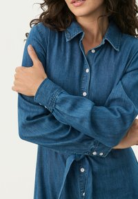 Denim košulja tamnoplave boje s ovratnikom, dugim rukavima s gumbima na manžetama i detaljem vezivanja u struku. Glatka tekstura s suptilnim sjajem.