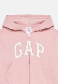 Haut à capuche zippé rose clair avec des poignets côtelés, des poches kangourou et le logo "GAP" embelli de strass. Tissu doux, design décontracté.