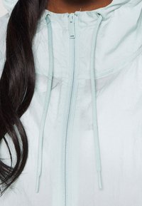 Långt mörkt hår som hänger över axeln på en person som bär en ljusblå zip-up jacka med huva och dragskor.