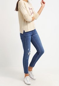 Beige Langarmoberteil mit rotem Sternmuster, kombiniert mit blauen Skinny Jeans mit abgenutzten Akzenten. Getragen mit weißen Turnschuhen.