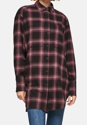 Kvinde iført langærmet oversized skjorte med knapper i mørk rosa og sort ternet mønster, kombineret med blå jeans.