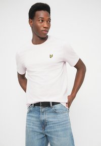 Világos rózsaszín pamut rövid ujjú póló kerek nyakkal, bal mellrészen egy kis sárga madár logóval. Látható kék farmernadrág és fekete öv.