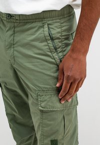 Olijfgroene cargobroek gemaakt van lichtgewicht stof, met een elastische tailleband, zij- en dijzakken, en zichtbare stikseldetails.