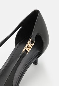 Chaussure à talons hauts synthétique noire avec un design découpé, arborant une accent en logo doré sur une sangle et une finition texturée.