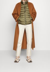 Persona vestida con un abrigo largo marrón, una chaqueta acolchada verde oliva, pantalones blanco sucio y zapatillas beige con acentos naranjas, de pie contra un fondo sencillo.